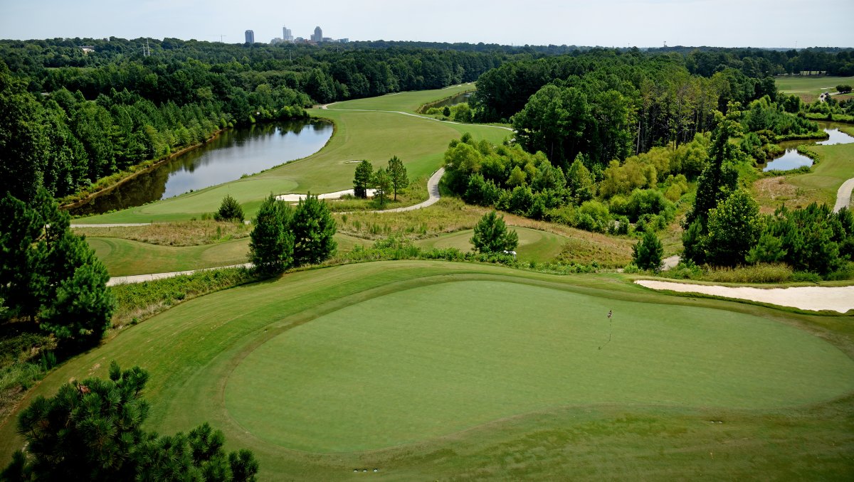 Aerial of green golf course surrounded by trees and Raleigh skyline in distance