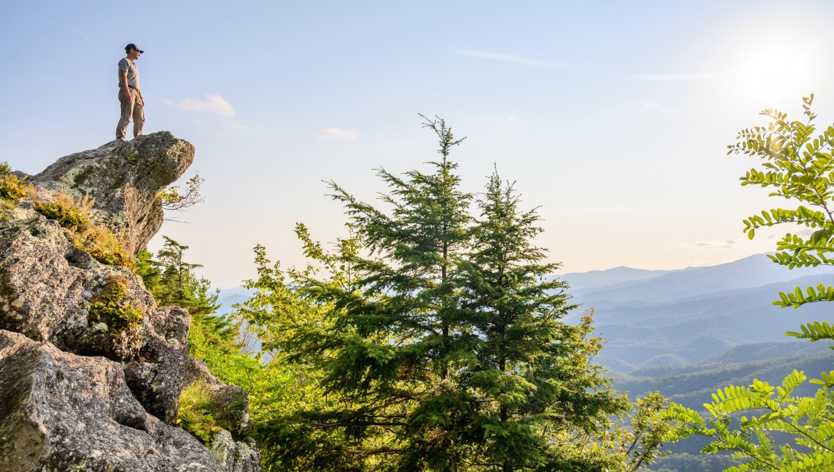 Man standing on top of The Blowing Rock outcropping surrounded by green trees and mountains in background