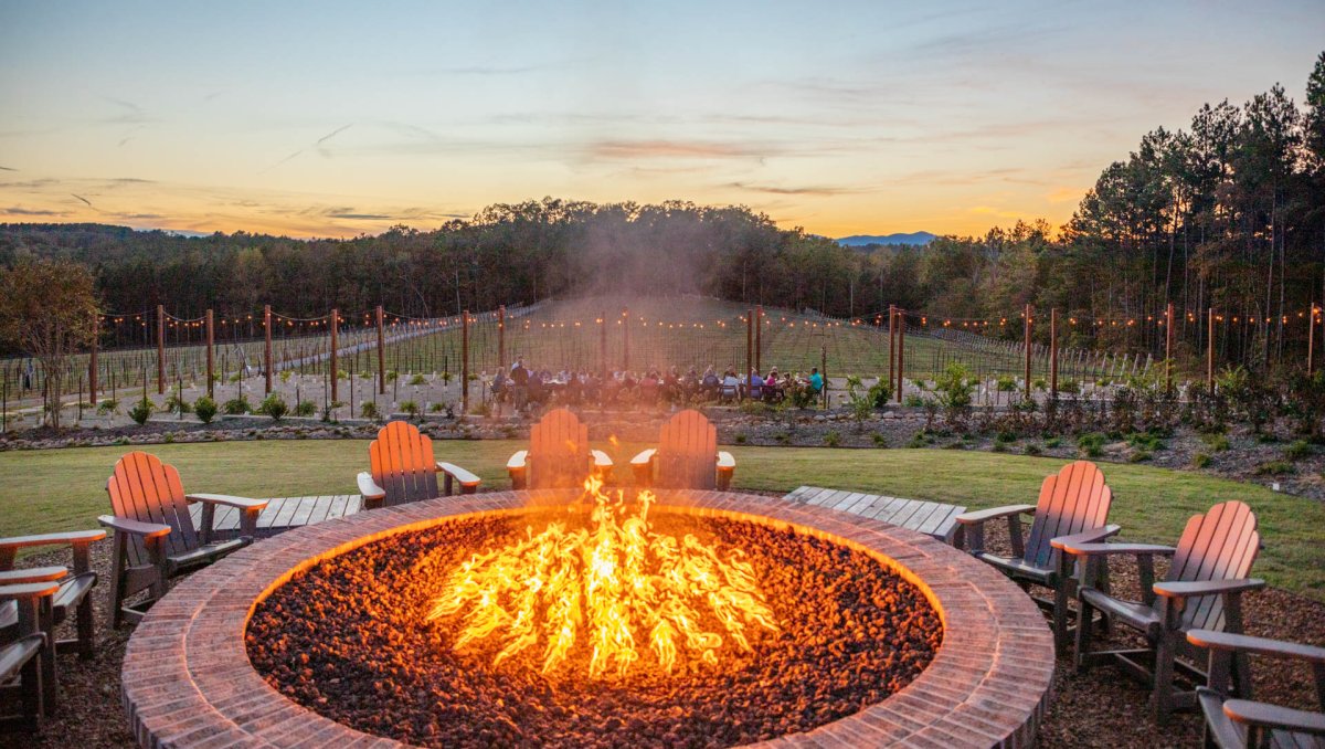 Huge fire in large fire pit surrounded by Adirondack chairs. Vineyard, trees and mountains in distance at sunset