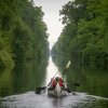Back of people in canoe paddling down canal, with green trees and foliage on either side during cloudy daytime