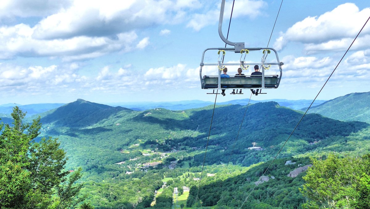 People riding scenic chairlift down mountain with mountains in background during summer