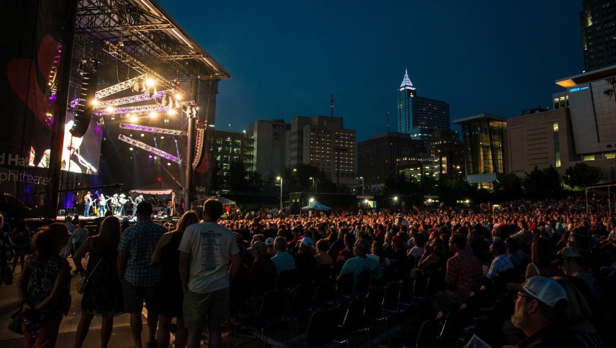 Wide view of huge crowd in front of band performing on stage with Raleigh skyline in background during nighttime