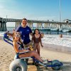 Woman in beach wheelchair smiling for camera with son and daughter in front of pier on beach during daytime