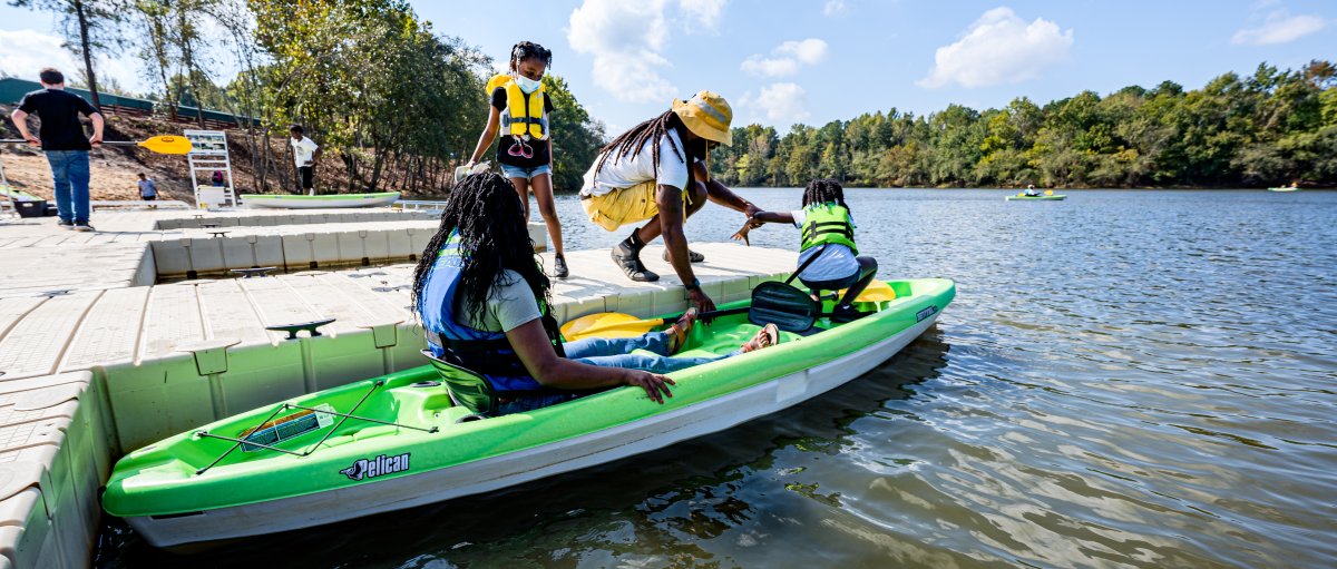 Person sitting in green kayak while an adult and child climb into front of kayak while parked at dock during daytime