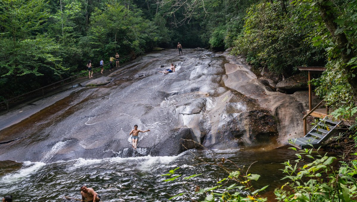 Kids sliding down rock into pool of natural water surrounded by green trees during daytime