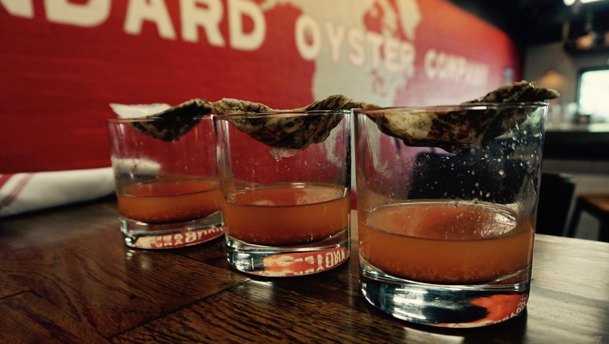 Three oyster shooters on wood table with oyster signage and red wall in background
