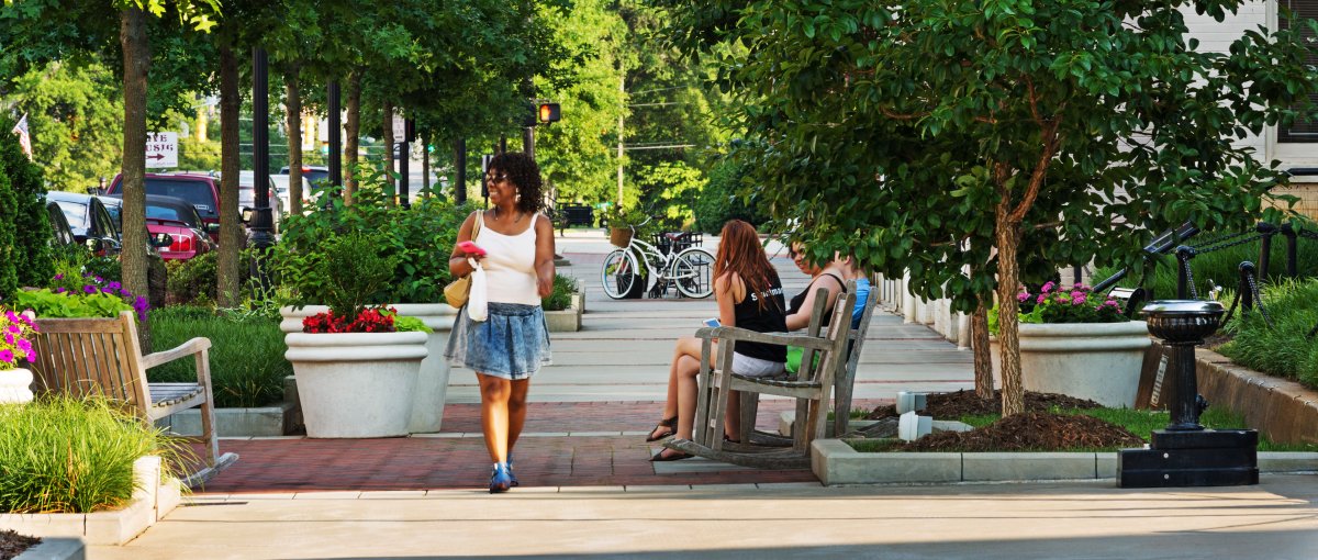 Woman walking down tree-lined streets of charming downtown with people sitting on rocking chairs on sidewalk