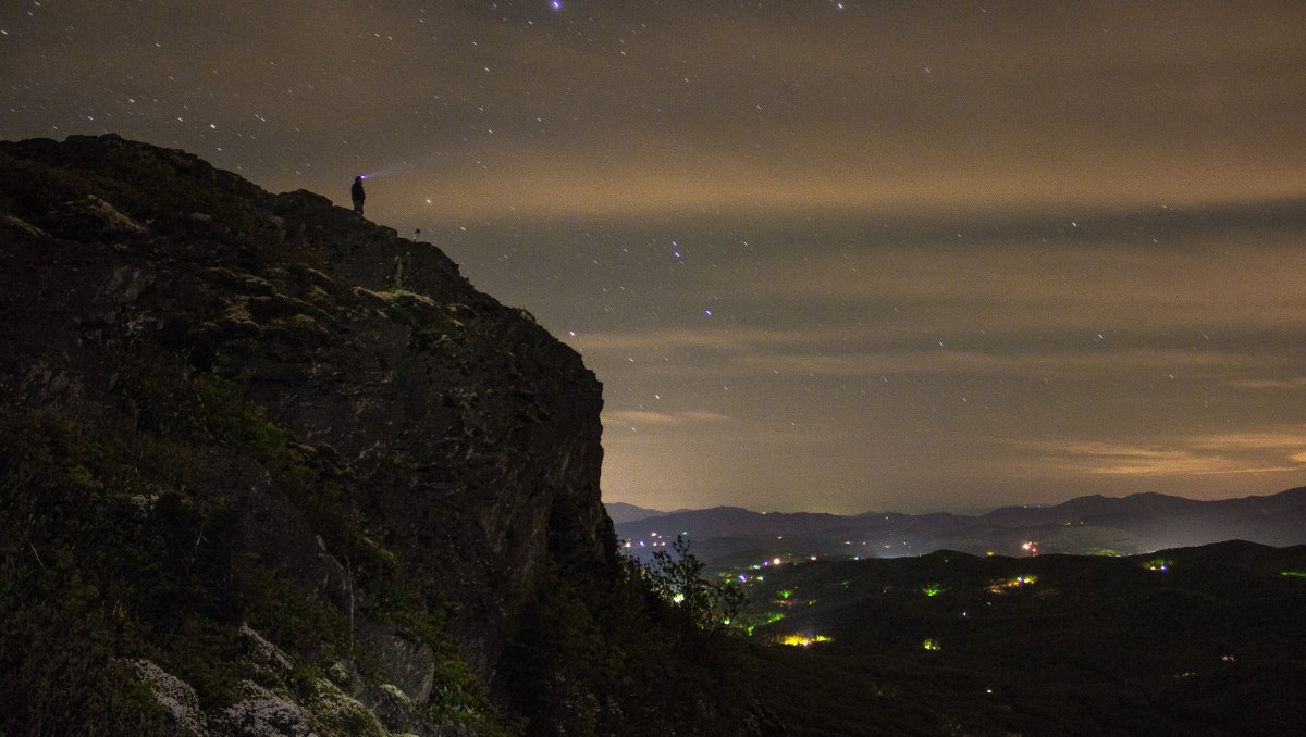 Person standing on mountain ridge at night with valley and starry sky
