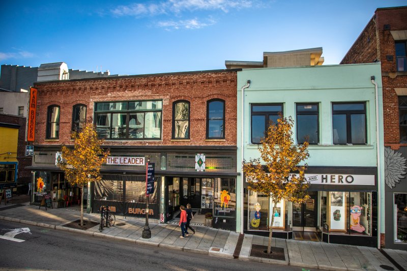 street scene with historic brick buildings as pedestrian walks along the sidewalk beneath the trees with autumn-colored leaves. featuring shops and eateries