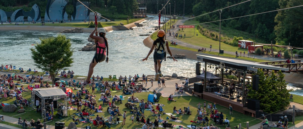 Two people going down zip lines with crowd of people on lawn below and whitewater river in background