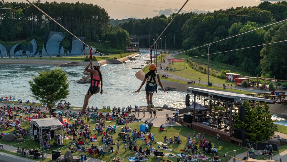 Two people going down zip lines with crowd of people on lawn below and whitewater river in background