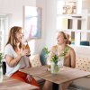 Two friends laughing while enjoying drink at table inside bright, airy restaurant.