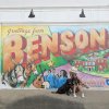 Woman and dog sitting in front of colorful mural reading "Greetings from Benson"