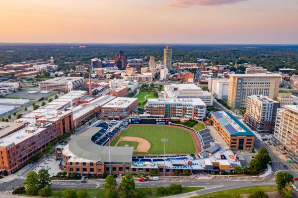 Aerial of Durham Bulls stadium surrounded by buildings with sun setting over Piedmont in background.