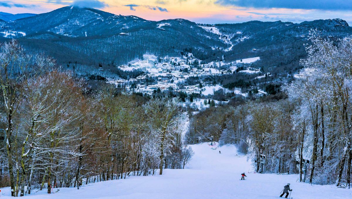 People skiing down hill with wintry trees and mountains during sunset in Sugar Mountain