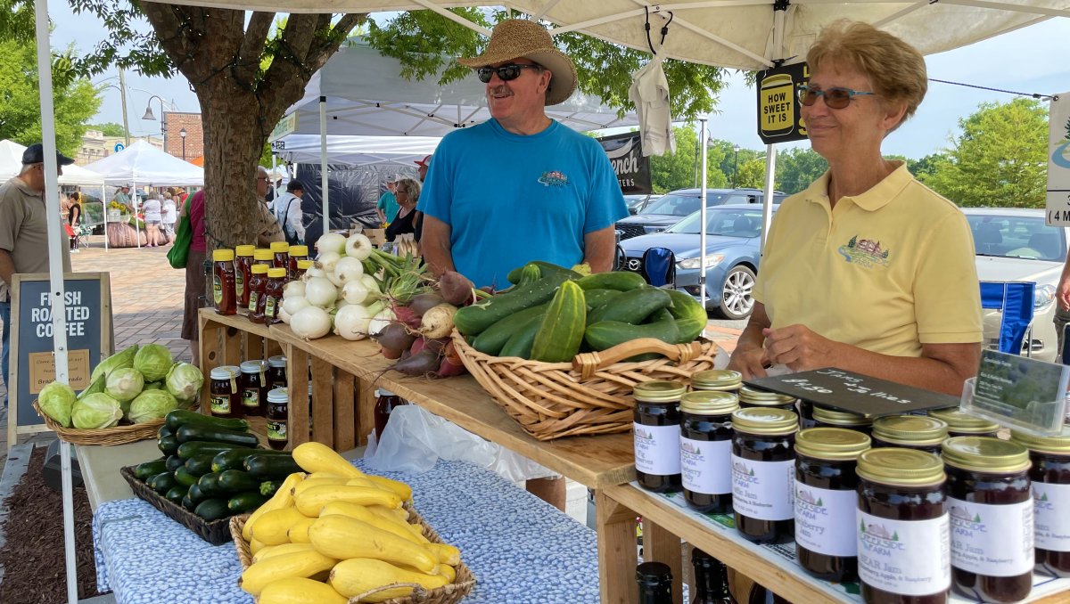 Two people standing at farmers market stand with veggies, jams and honey on tables in front of them