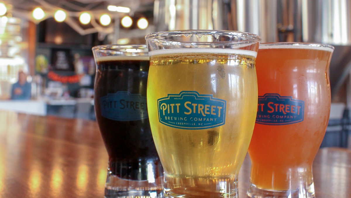 Closeup of three types of beers from Pitt Street Brewing with logos on mugs and patrons and brewery in background