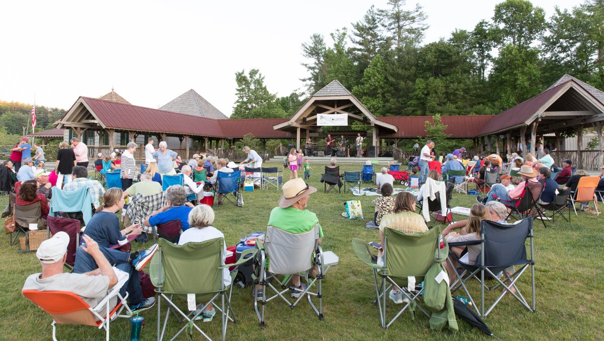 People sitting on lawn enjoying live music from pavilion during daytime.