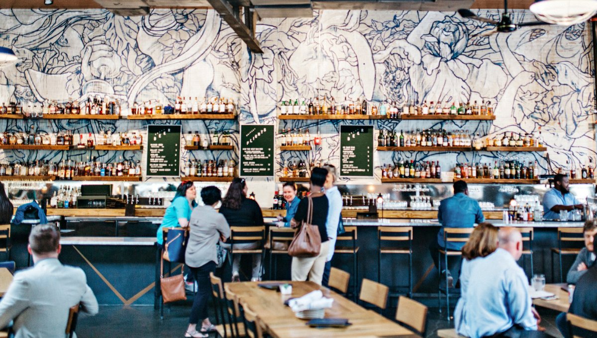 Interior of trendy bar and restaurant with people sitting at tables and bar with floral wallpaper on back wall