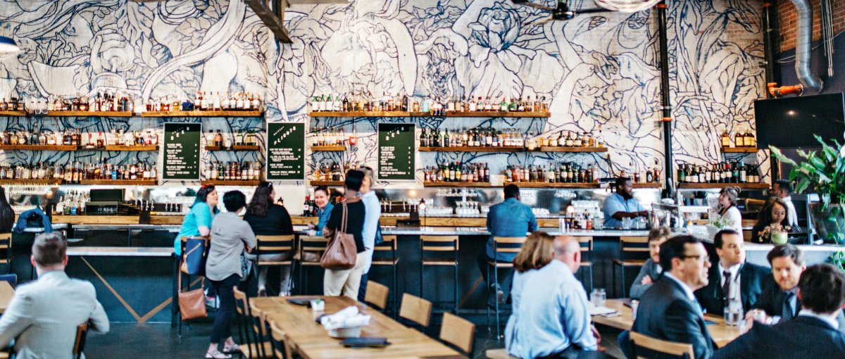 Interior of trendy bar and restaurant with people sitting at tables and bar with floral wallpaper on back wall