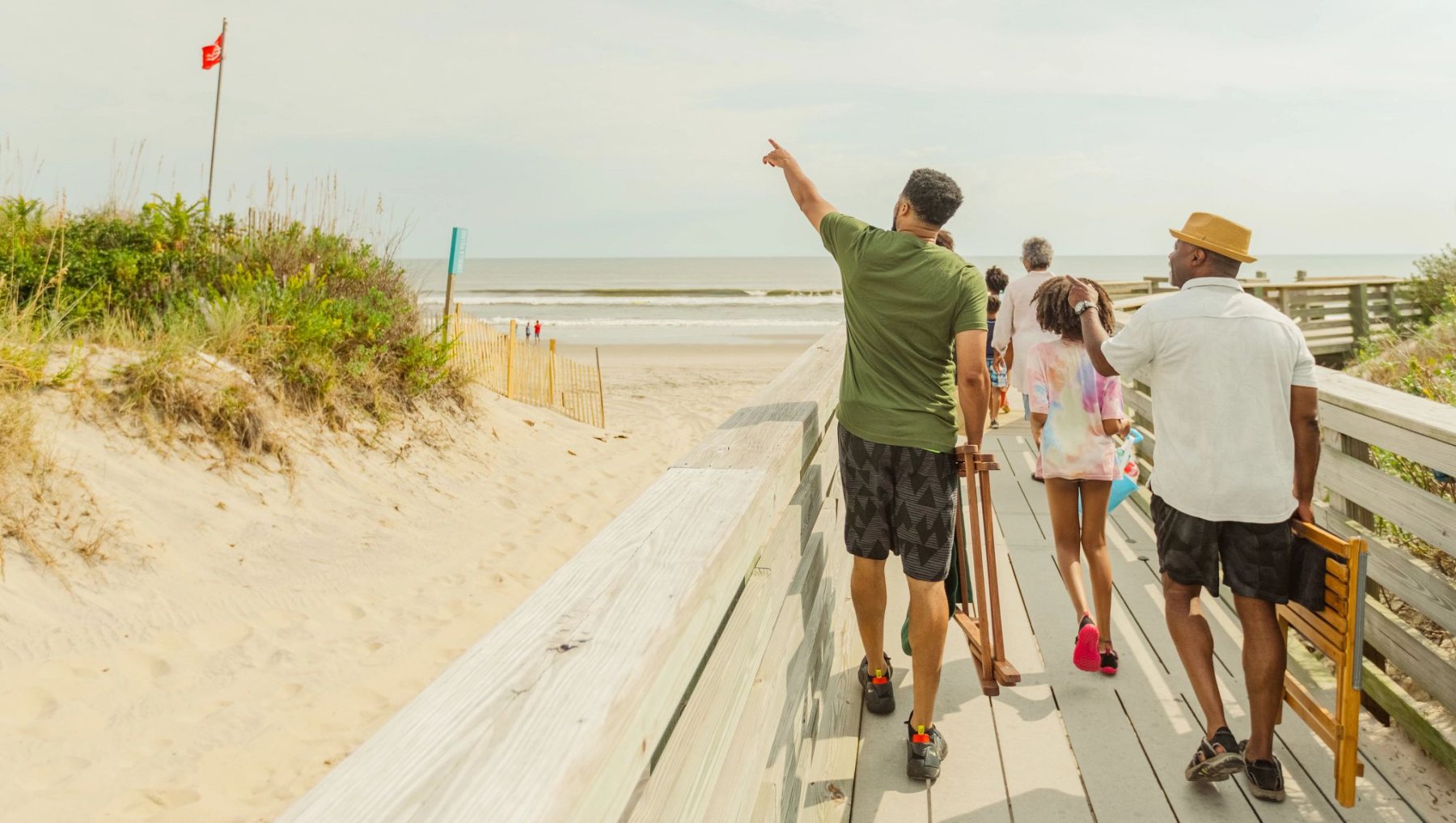 Wide landscape view of family carrying beach chairs and walking along dock at Corolla Beach in the Outer Banks in daytime with focal on men pointing to ocean horizon blurred in background.