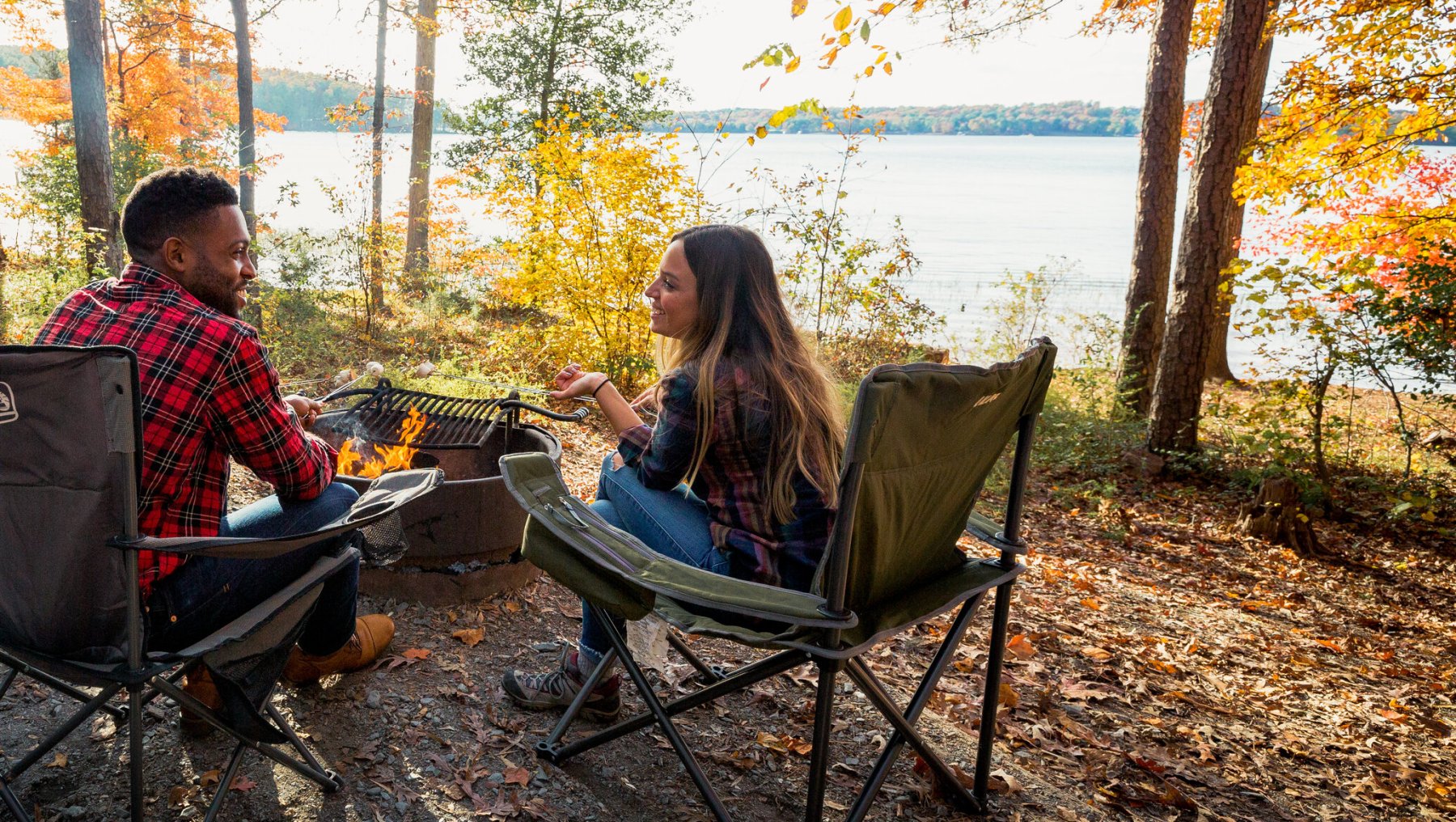 Three friends sitting around campfire at campsite bordering lake, surrounded by fall foliage