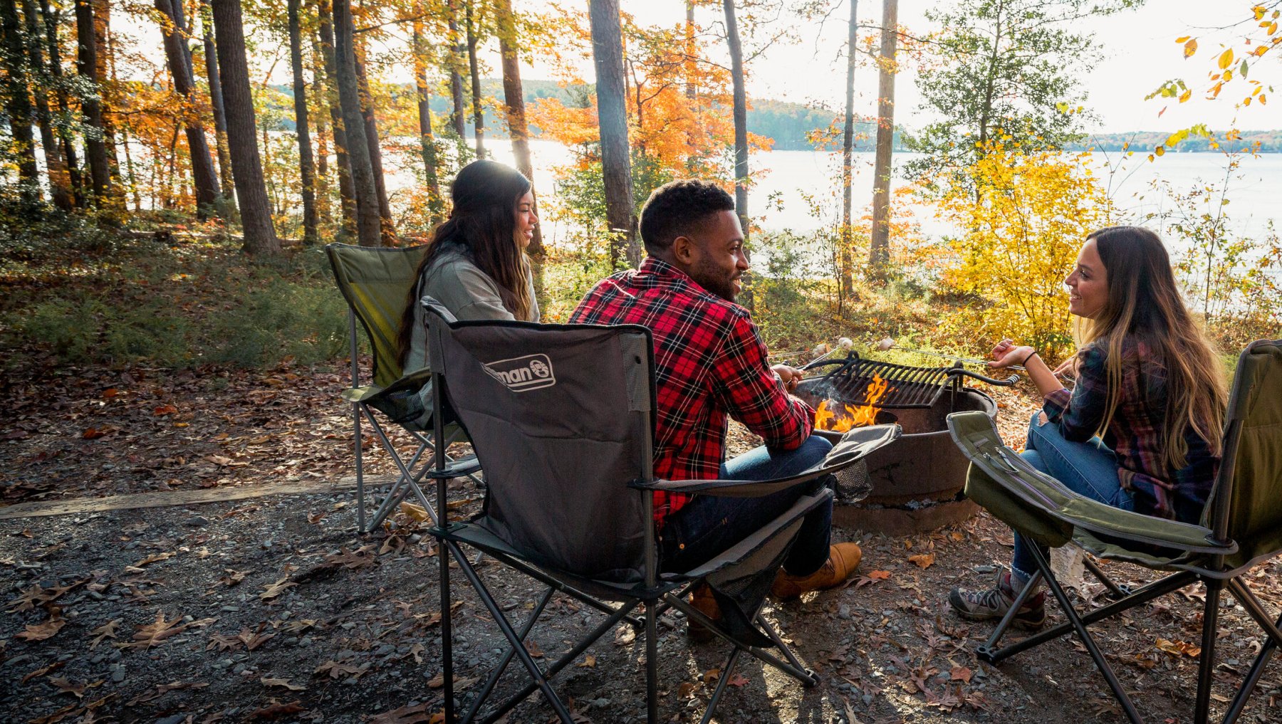 Three friends sitting around campfire at campsite bordering lake, surrounded by fall foliage