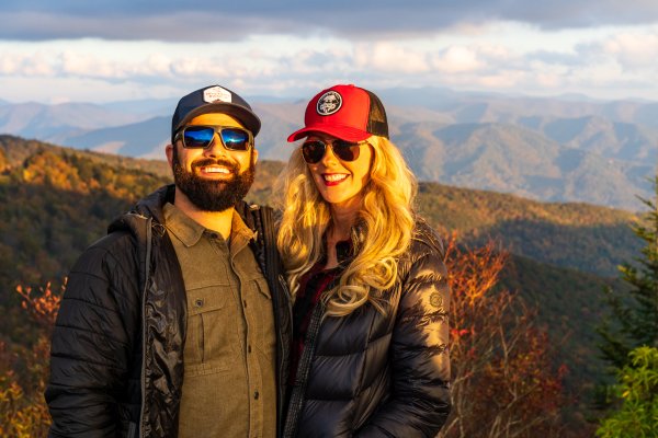 Couple wearing jackets and hats smiling at camera in front of scenic mountains during daytime
