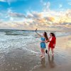 Two women taking a selfie on beach near ocean under blue and orange sky