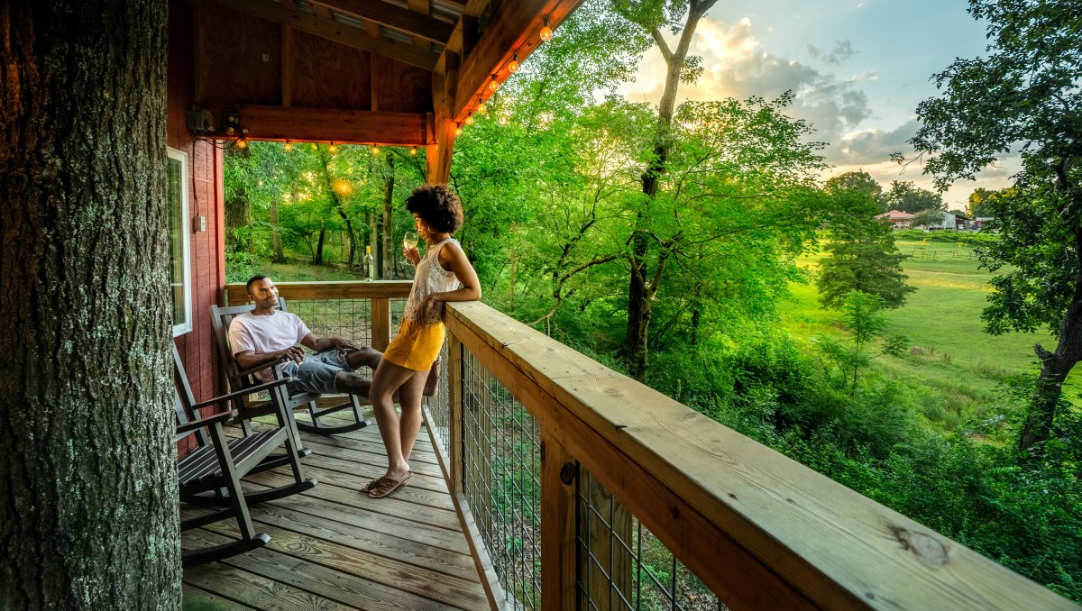 Couple enjoying wine on treehouse balcony with green trees and grounds to the right