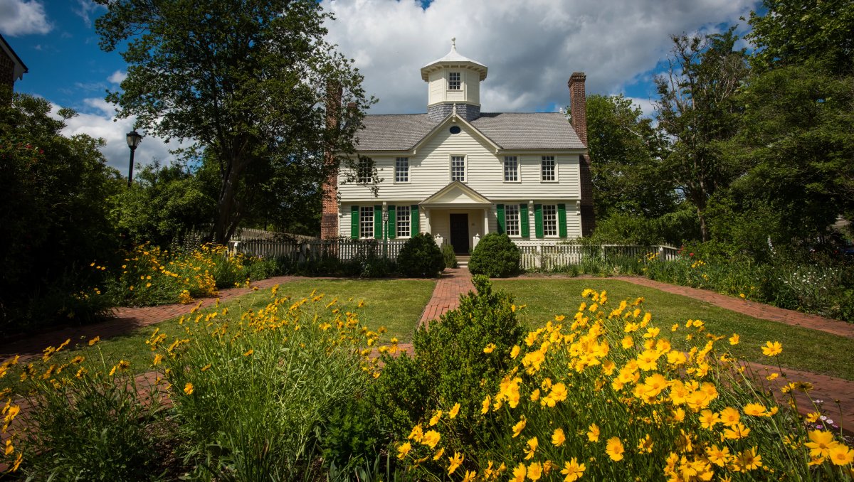 Exterior of historic house with flowers and gardens in foreground and green foliage on each side during semicloudy daytime