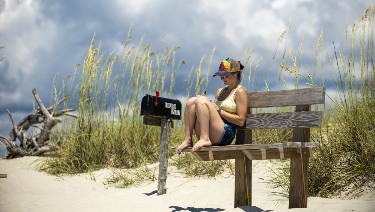 Woman sitting on bench near Kindred Spirit Mailbox with beach oats in background
