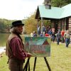 Man displaying painting of log cabin as people walk in and out of cabin in background during fall day
