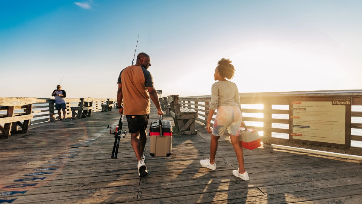 Man and child holding fishing gear and walking down fishing pier under blue sky during daytime