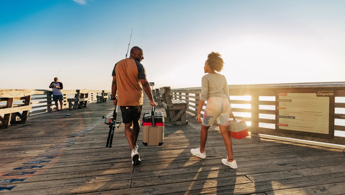 Man and child holding fishing gear and walking down fishing pier under blue sky during daytime