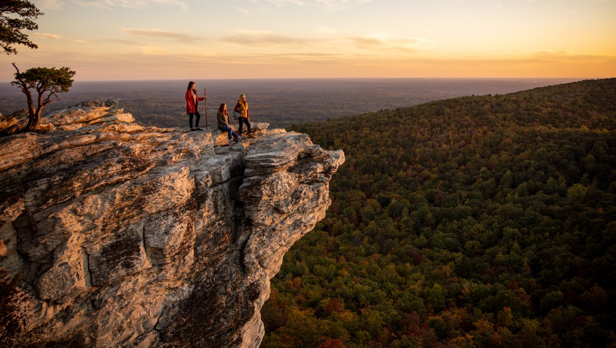 Three people on rock ledge looking out over fall foliage and views during sunset