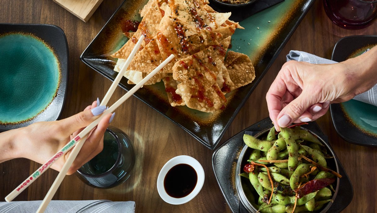 Overhead of Asian-influenced food in middle of table with hands reaching for the food