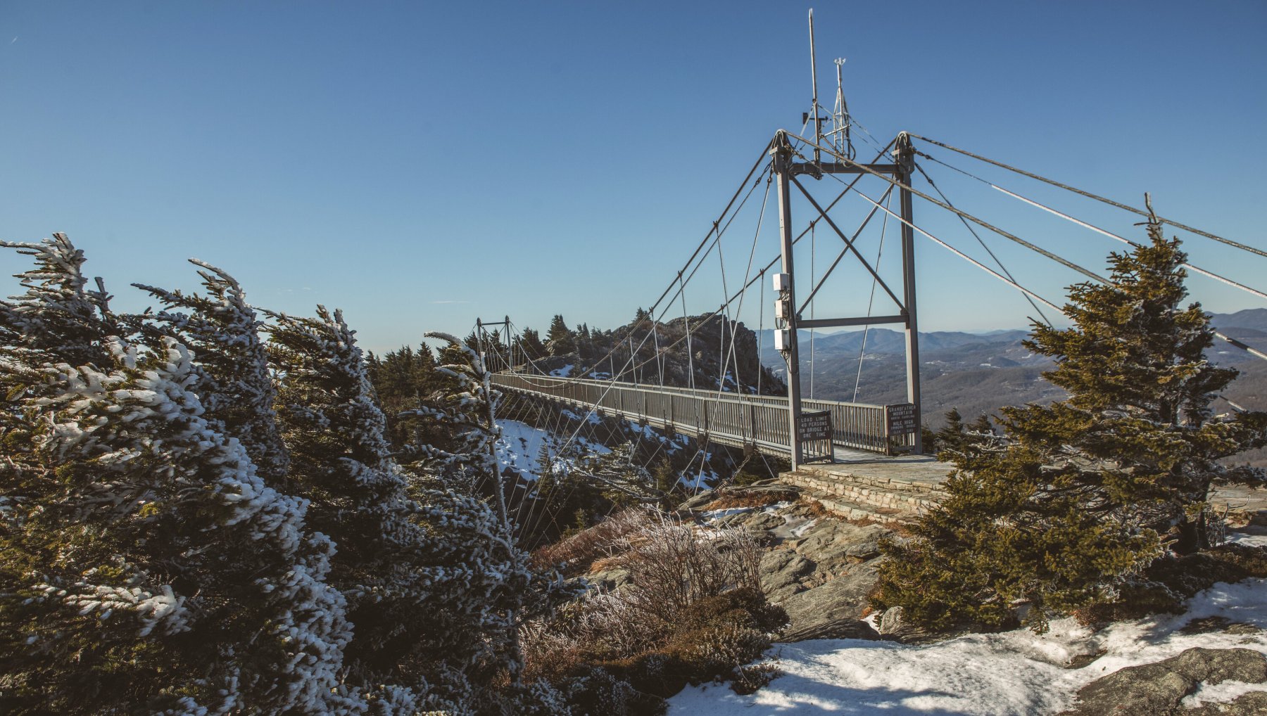 Suspension bridge at top of mountain extending to other side of mountain, surrounded by snowy evergreen trees during daytime