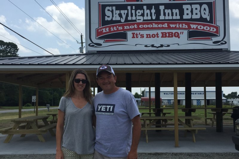 Man and woman smiling for camera in front of sign reading Skylight Inn BBQ