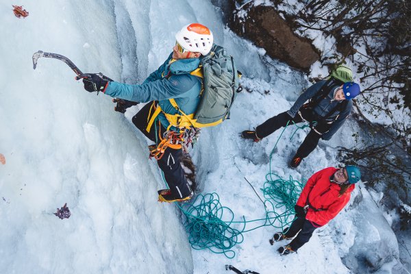 Man beginning to ice climb up wall of ice with people on ground watching him.