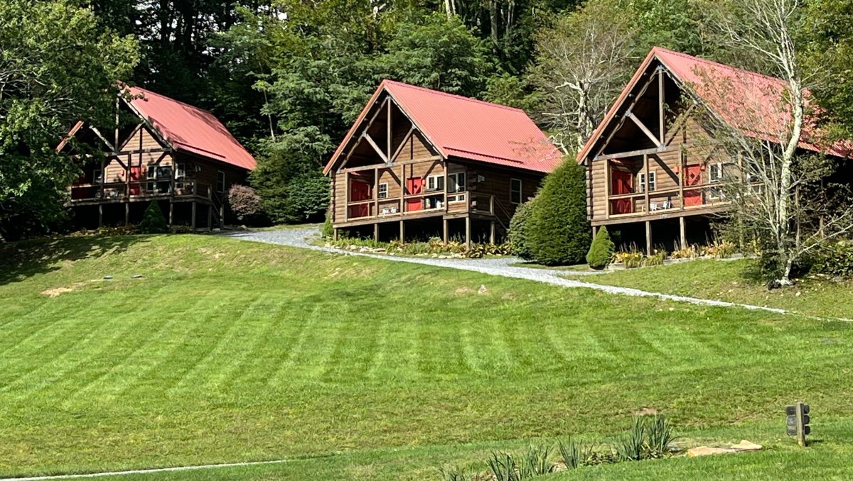 Exterior of three charming mountain cabins sitting on hill overlooking expansive green lawn.