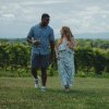 Couple smiling at each other and holding wine as they walk around vineyard