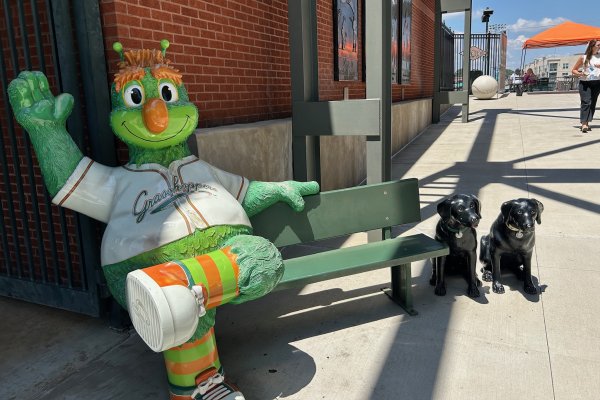 Guilford the Grasshopper mascot sitting on bench and waving at camera at baseball game.