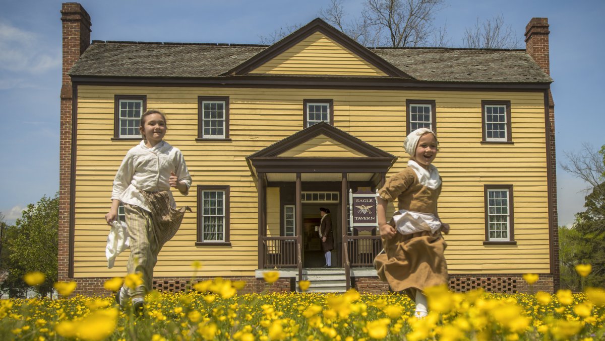 Two children dressed in 1700s period garb running in field of yellow flowers in front of historic building