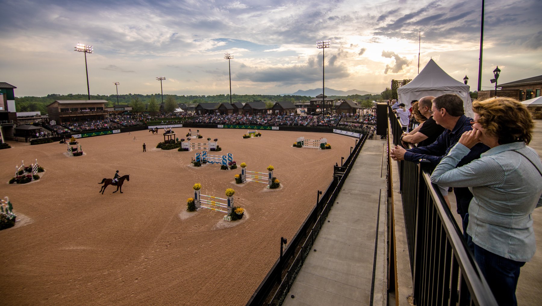 People watching horse show at equestrian center with mountains in distance.