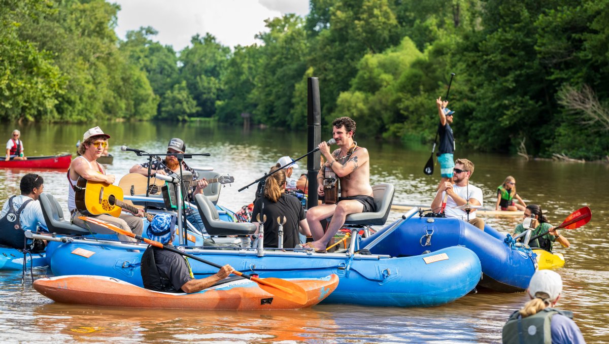 Band performing on river raft with people in kayaks surrounding them during on-water concert
