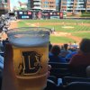 Person holding Durham Bulls-branded beer in stands with baseball game playing in background.