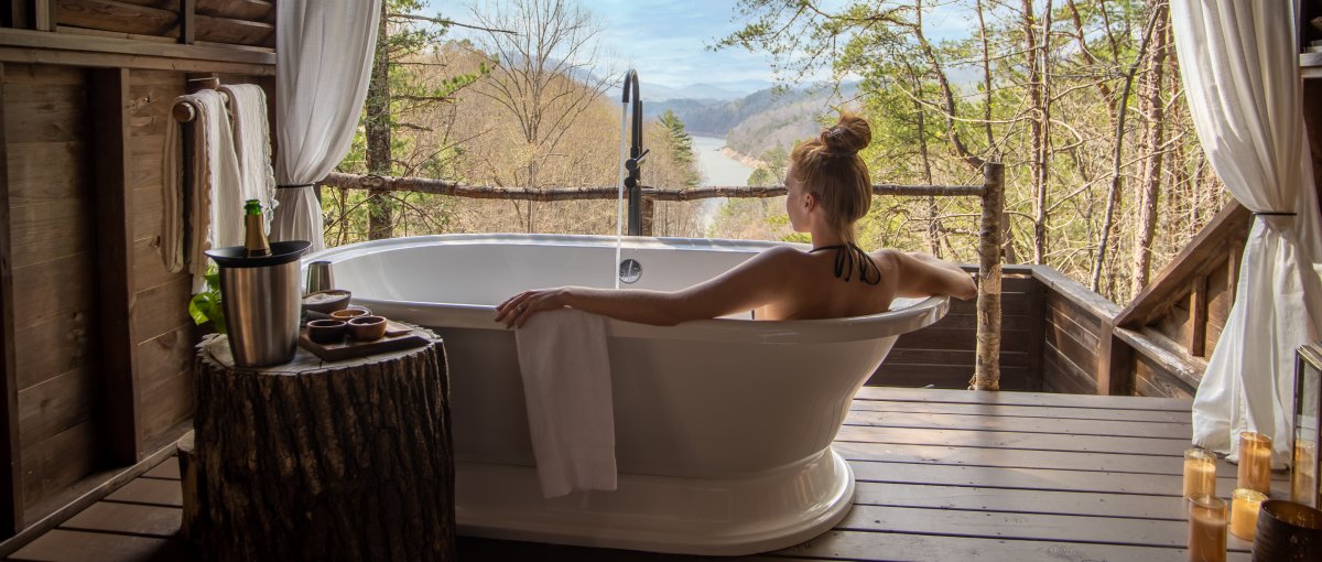 Woman in soaking tub in treehouse with forest, mountains and river in distance