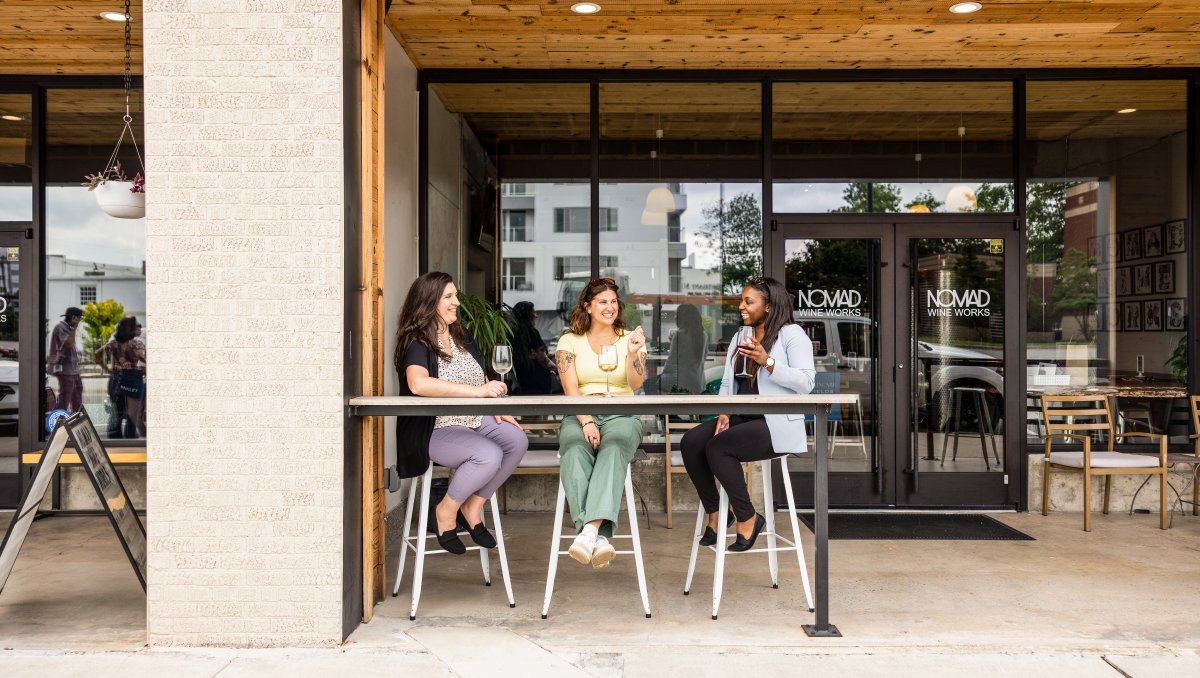 Three smiling friends drinking wine on front patio of wine store during daytime.