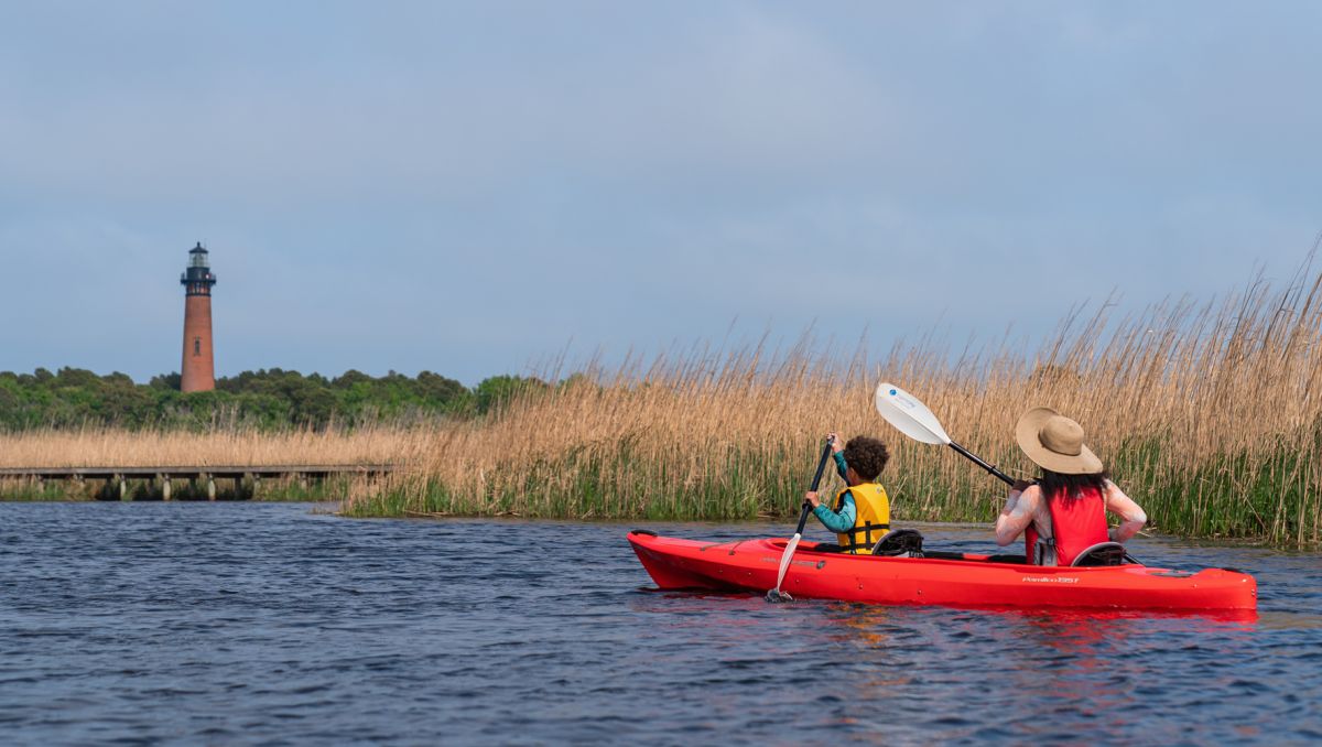 Woman and child in red kayak paddling through march with lighthouse in background